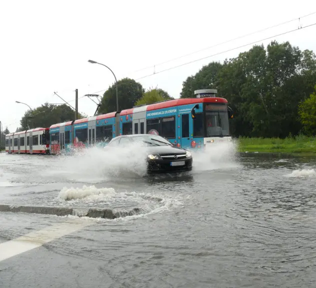 Ein dunkles Auto, das neben einer Stadtbahn über eine geflutete Straße fährt. Das Wasser spritzt.