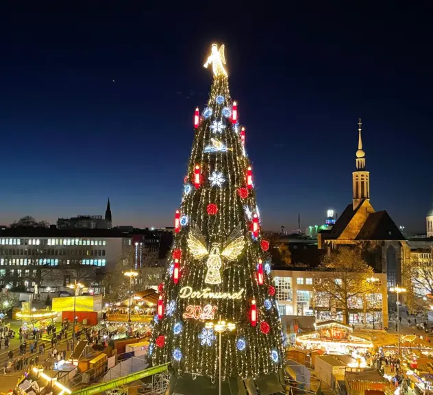 Der Weihnachtsbaum der Dortmunder Weihnachtsstadt im Jahr 2021. Er ist mit verschiedenen Lichtern beleuchtet und um ihn herum sind beleuchtete Stände.