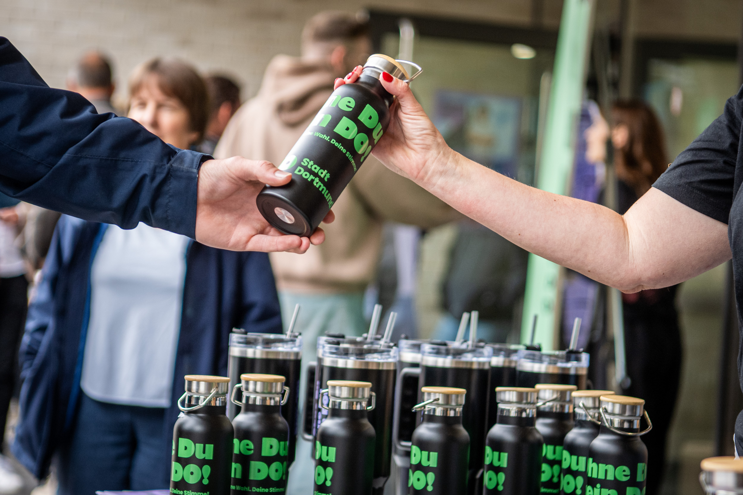 Nahaufnahme von einer Person, die einer anderen Person eine "ohne du kein do" Trinkflasche überreicht