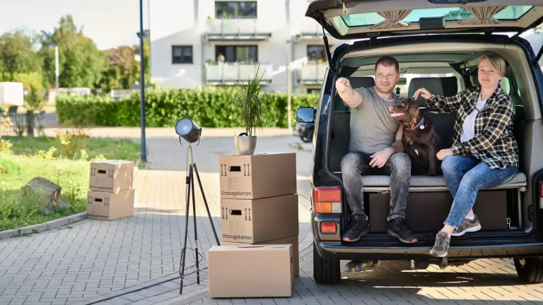 Ein Paar sitzt mit einem braunen Hund in einem Van, davor stehen Umzugskartons