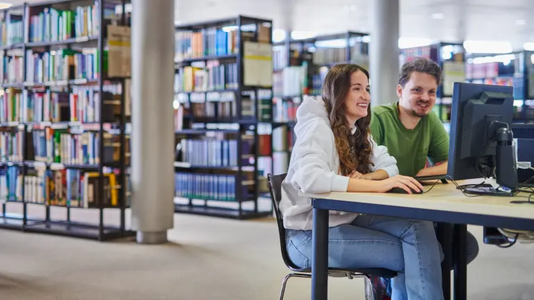 Zwei Personen sitzen an einem PC der Stadt- und Landesbibliothek. Im Hintergrund stehen Bücherregale