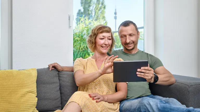 Paar sitzt auf einer Couch und sieht sich etwas auf einem Tablet an. Im Hintergrund sieht man durch ein Fenster den Florianturm.