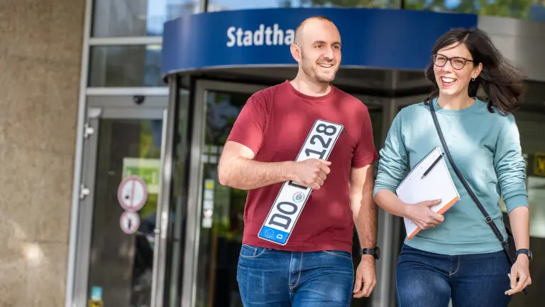Ein Mann und eine Frau verlassen lächelnd mit einem neuen Kennzeichen in der Hand ein Gebäude der Stadt Dortmund