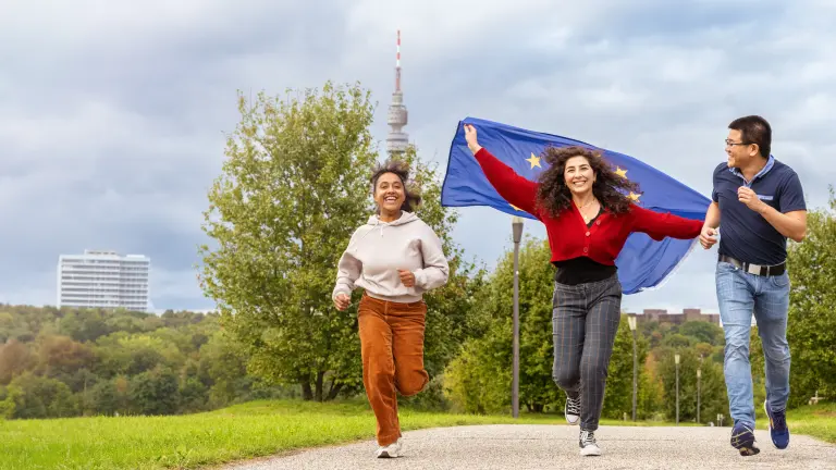 Drei Personen laufen mit Europa-Flagge über einen Weg. Im Hintergrund sieht man hinter Bäumen Dortmunder Wahrzeichen