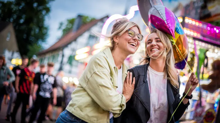 Zwei Frauen laufen lachend über die Kirmes in Dortmund. Eine Person hält einen Luftballon in der Hand.