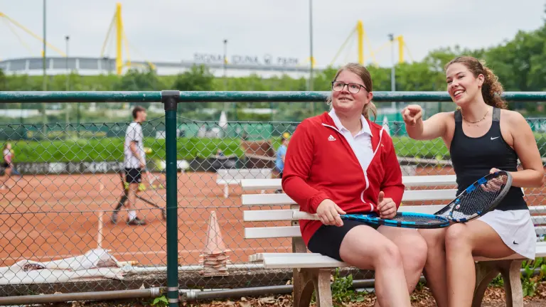 Zwei Frauen sitzen mit Tennisschlägern in der Hand auf einer Bank. Im Hintergrund sieht man einen Tennisplatz auf dem Tennis gespielt wird und den Signal Iduna Park
