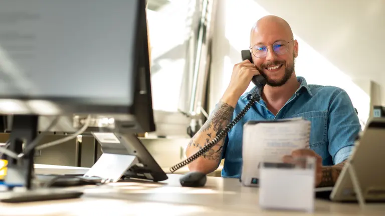 Mann sitzt lächelnd im Büro mit Telefon und Notizblock in der Hand