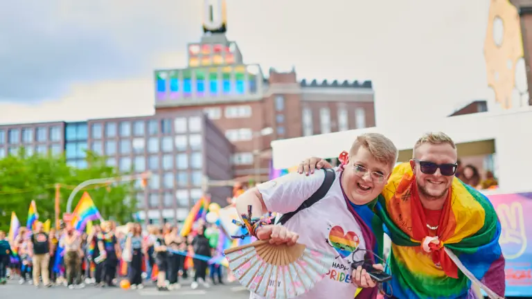 Zwei Personen lachen auf einer LGBTQIA-Veranstaltung in die Kamera. Im Hintergrund zeigt der Dortmunder U-Turm die LGBTQIA-Flagge.