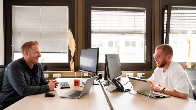Zwei Männer sitzen einander gegenüber an zwei Schreibtischen in einem Büro und lachen