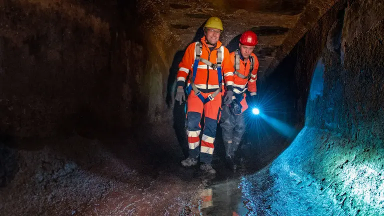 Zwei Männer in orangener Arbeitskleidung stehen mit Taschenlampe in einem Abwasserkanal
