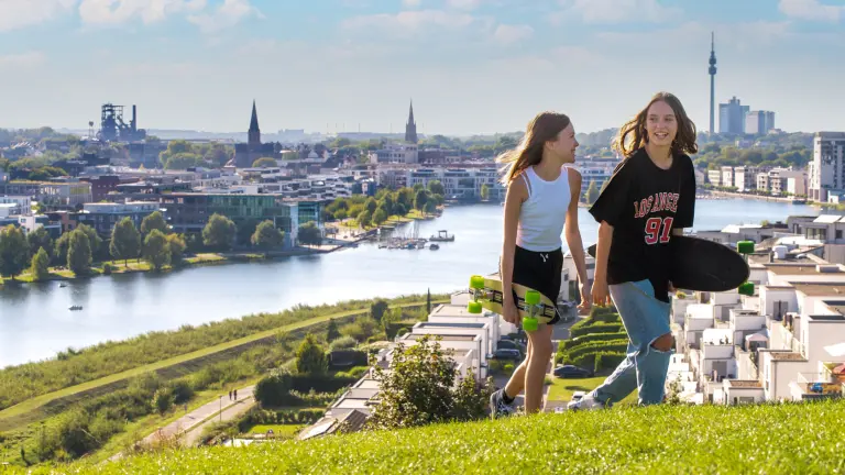 Zwei Mädchen spazieren mit ihren Skateboards unter dem Arm über einen Hügel, im Hintergrund ist der Phönixsee zu sehen