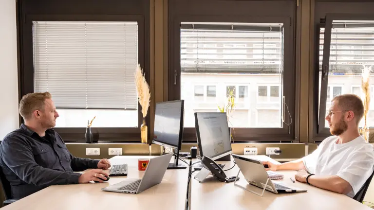 Zwei Männer sitzen einander gegenüber im Büro und arbeiten jeweils an ihrem Schreibtisch