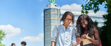 Zwei junge Menschen schauen freudig auf ein Smartphone, im Hintergrund ist der Mathematik-Turm der TU Dortmund.