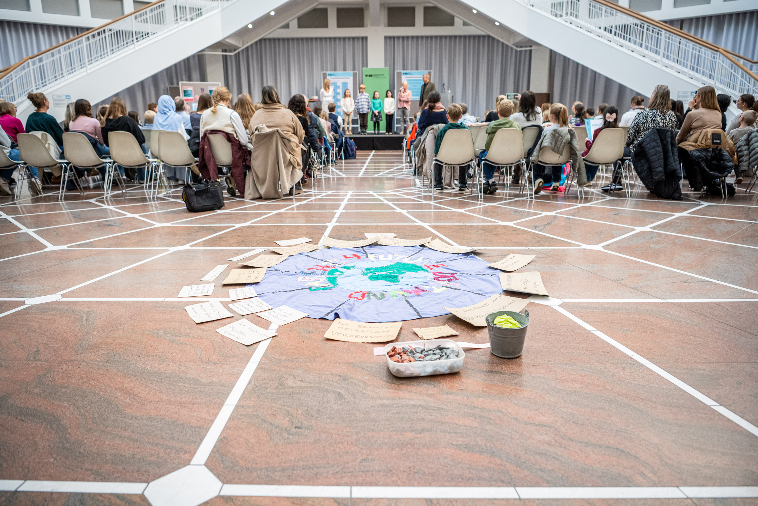 Im Vordergrund Zettel und Gebasteltes auf dem Boden, im Hintergrund die Preisverleihung zum Preis Umweltbewusste Schule 2024