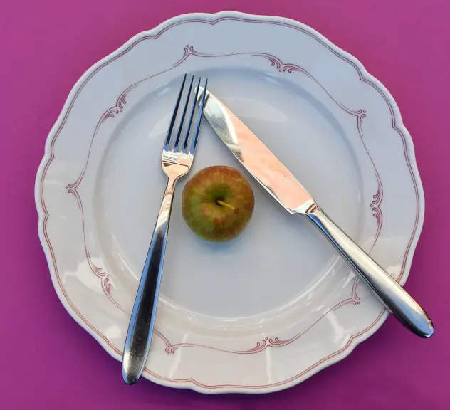 Gabel und Messer gekreuzt auf einem Teller, auf dem ein kleiner Apfel liegt
