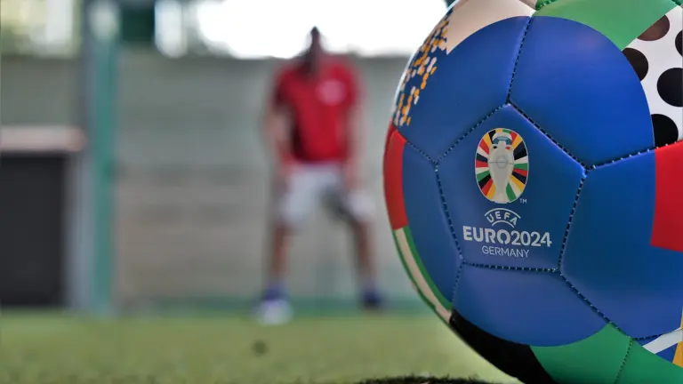 Im Vordergrund liegt der EM-Ball der Fußballeuropameisterschaft 2024, im Hintergrund steht ein Torwart im Tor bereit.