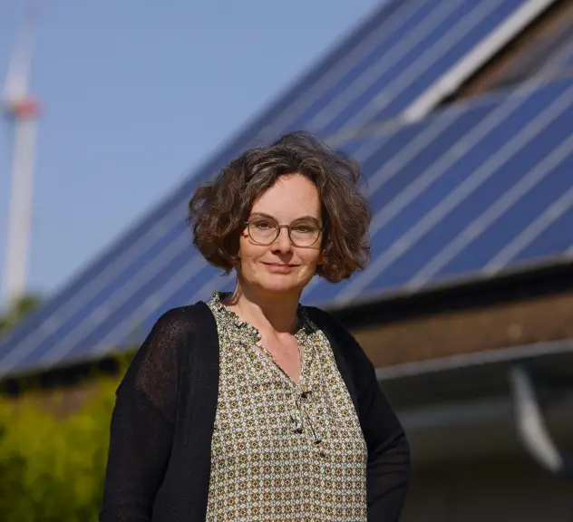 Eine Frau steht im Vordergrund, hinter ihr ist das Dach eines Hauses, auf dem eine Photovoltaikanlage angebracht ist. Ganz im Hintergrund steht ein Windkraftwerk.