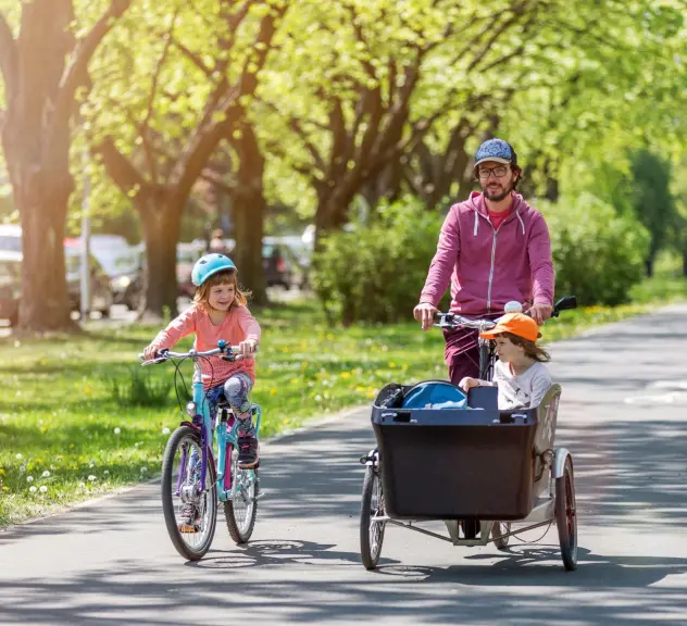 Mann fährt mit einem Lastenfahrrad, in dem ein Kind sitzt. Daneben fährt auf einem Fahrrad ein größeres Kind.