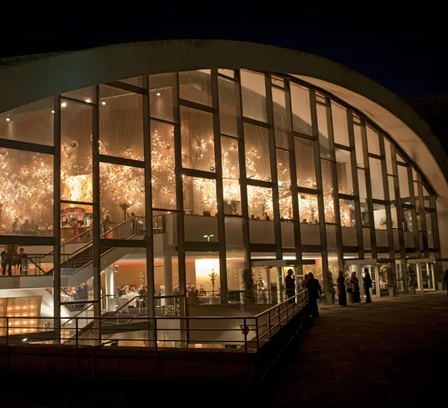 Das beleuchtete Opernhaus Dortmund bei Nacht, drinen hinter der Glasfassade und draußen stehen Menschen.