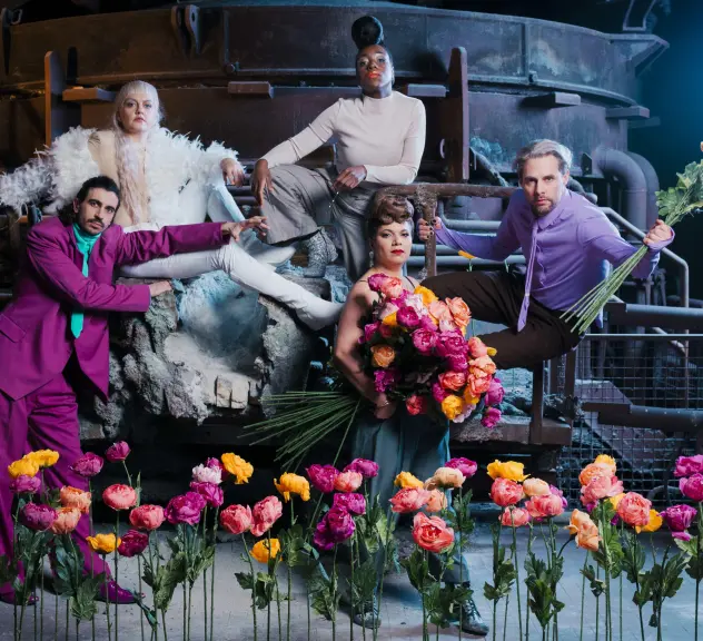 Promofotografie der Darstellerinnen und Darsteller der Inszenierung "Bakchen". Die Schauspieler tragen imposante Kostüme, vor ihnen ragt ein Blumenmeer als langstieligen Rosen