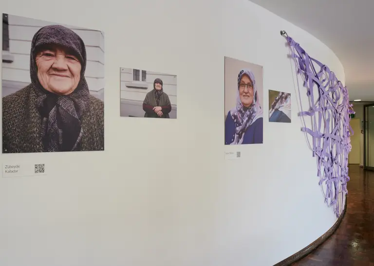 Wandrundung, an der Fotos in unterschiedlicher Größe von verschiedenen Frauen mit Kopftüchern hängen. Unter den Fotos befinden sich URL-Codes. Weiter hinten an der Wand ist ein netzähnliches Konstrukt aus violetten Schnüren angebracht.