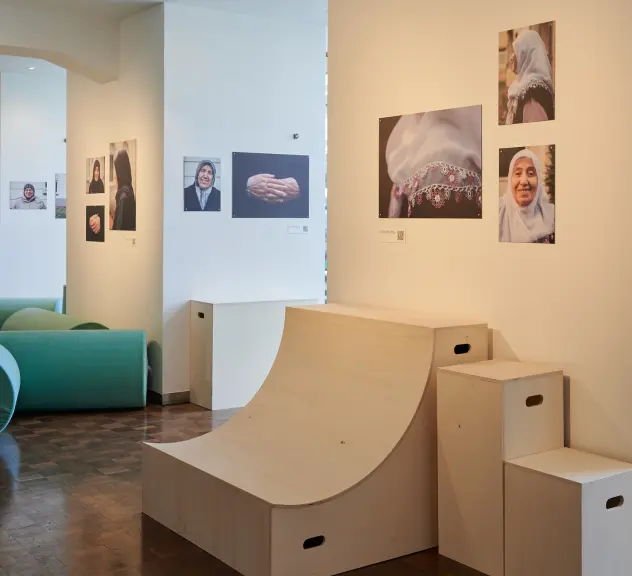 Ausschnitt eines Raumes, Im Raum stehen verschiedenförmige Kisten und aus Holz und Objekte aus grünem Kunststoff. An den Wänden hängen Fotos von verschiedenen kopftuchtragenden Frauen.
