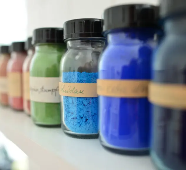 verschiedene bunte Pigmentfarben stehen in kleinen beschrifteten Glasflaschen aufgereiht auf einem Regal