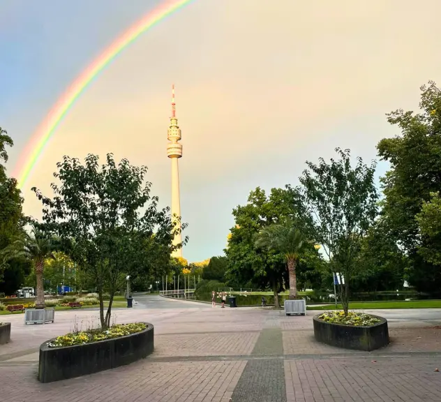 Ein beeindruckender Regenbogen spannt sich über den Westfalenpark in Dortmund mit Blick auf den Fernsehturm, umgeben von üppigem Grün und gepflegten Wegen, symbolisiert ein perfektes Ausflugsziel für Natur- und Freizeitliebhaber.*innen