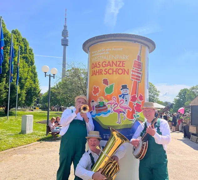 Drei musikalische Gärtner spielen Trompete, Tuba und Banjo vor einem bunten Werbepfeiler für den Westfalenpark, mit dem Florianturm im Hintergrund, während des sonnigen Frühlingsmarkts