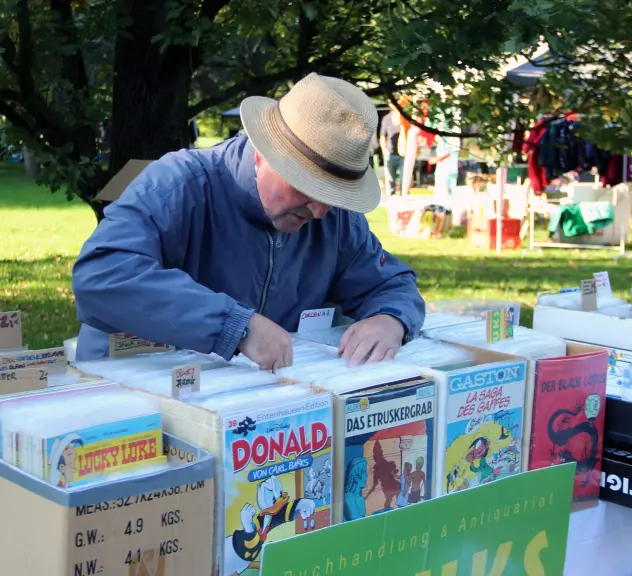 Ein Mann mittleren Alters mit Strohhut durchstöbert Comic-Sammlungen auf dem Trödelmarkt im Westfalenpark, darunter Ausgaben von Lucky Luke und Donald Duck.