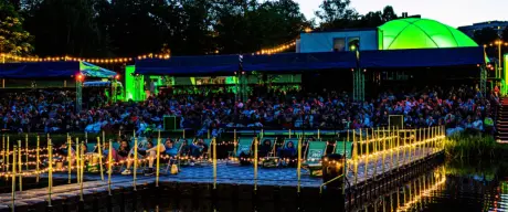 Besucher*nnen genießen das PSD Bank Sommerkino im Westfalenpark bei Nacht, mit bunter Beleuchtung und Spiegelung im Wasser