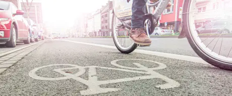 Fahrrad(fahrer*in) fährt auf einem Radweg. Fahrradsymbol in weiß auf dem Boden des Radweges. Im Hintergrund parkende Autos.