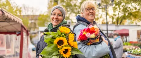 Zwei Frauen mit Blumensträußen im Arm stehen Rücken an Rücken auf dem Dortmunder Markt und lachen in die Kamera