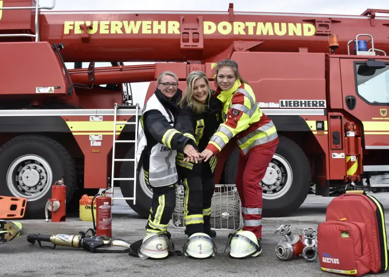 Drei Personen in Feuerwehruniform halten die Hände zusammen und stehen vor einem Feuerwehrwagen