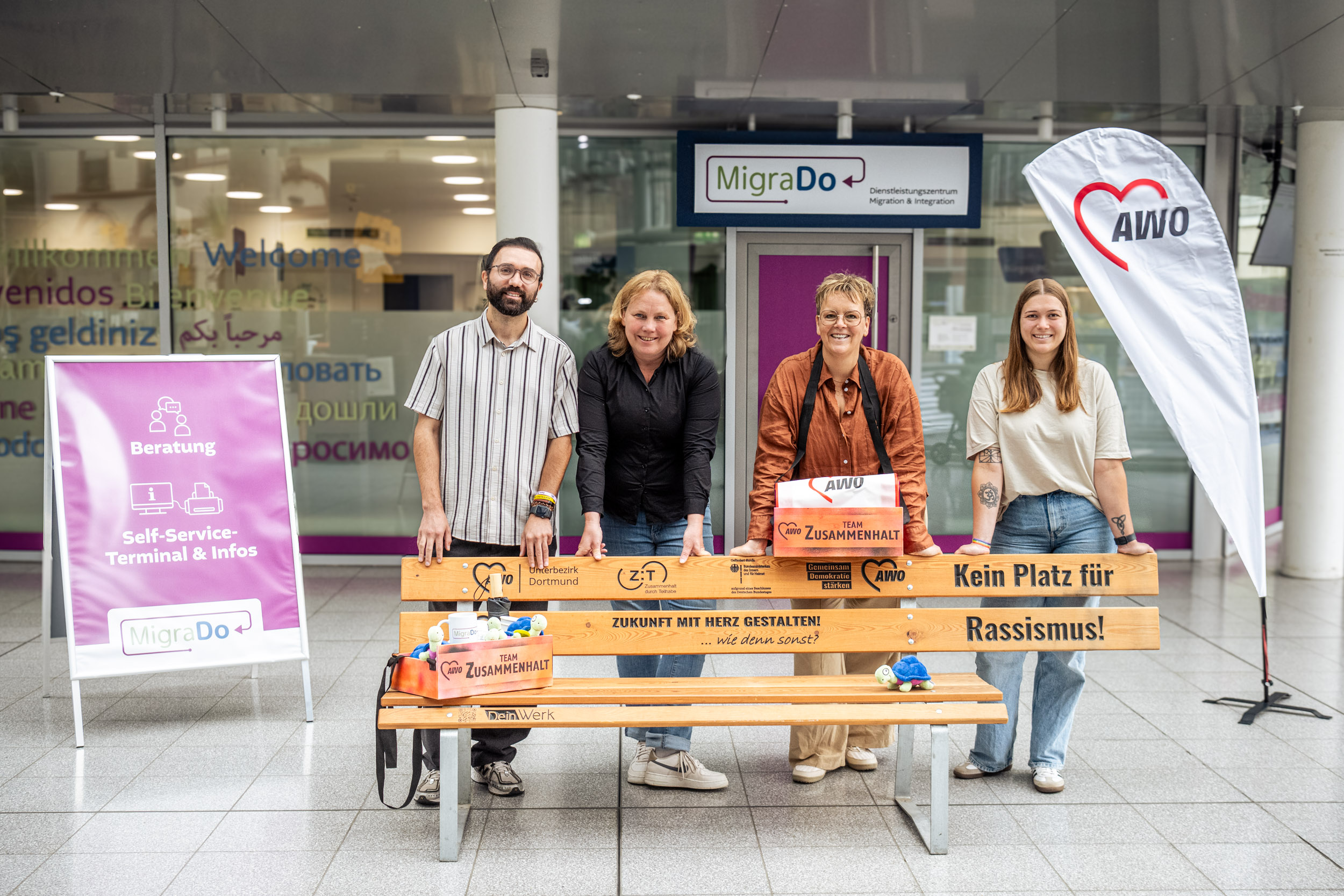 Vier Personen stehen hinter einer Holzbank, auf der steht: "Kein Platz für Rassismus!". Die Sitzfläche unter diesem Spruch ist ausgespart. 