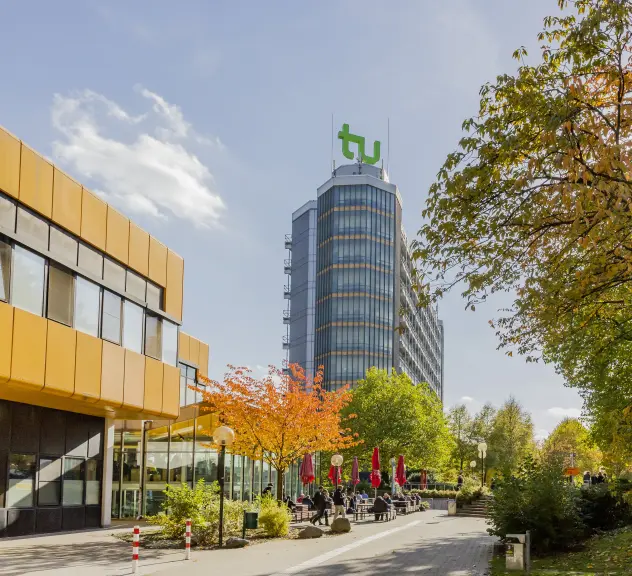 Mathetower der Technischen Universität Dortmund mit dem großen TU Logo. Außerdem Ausschnitt der Terrasse der Cafeteria mit Menschen, die in der Sonne sitzen.