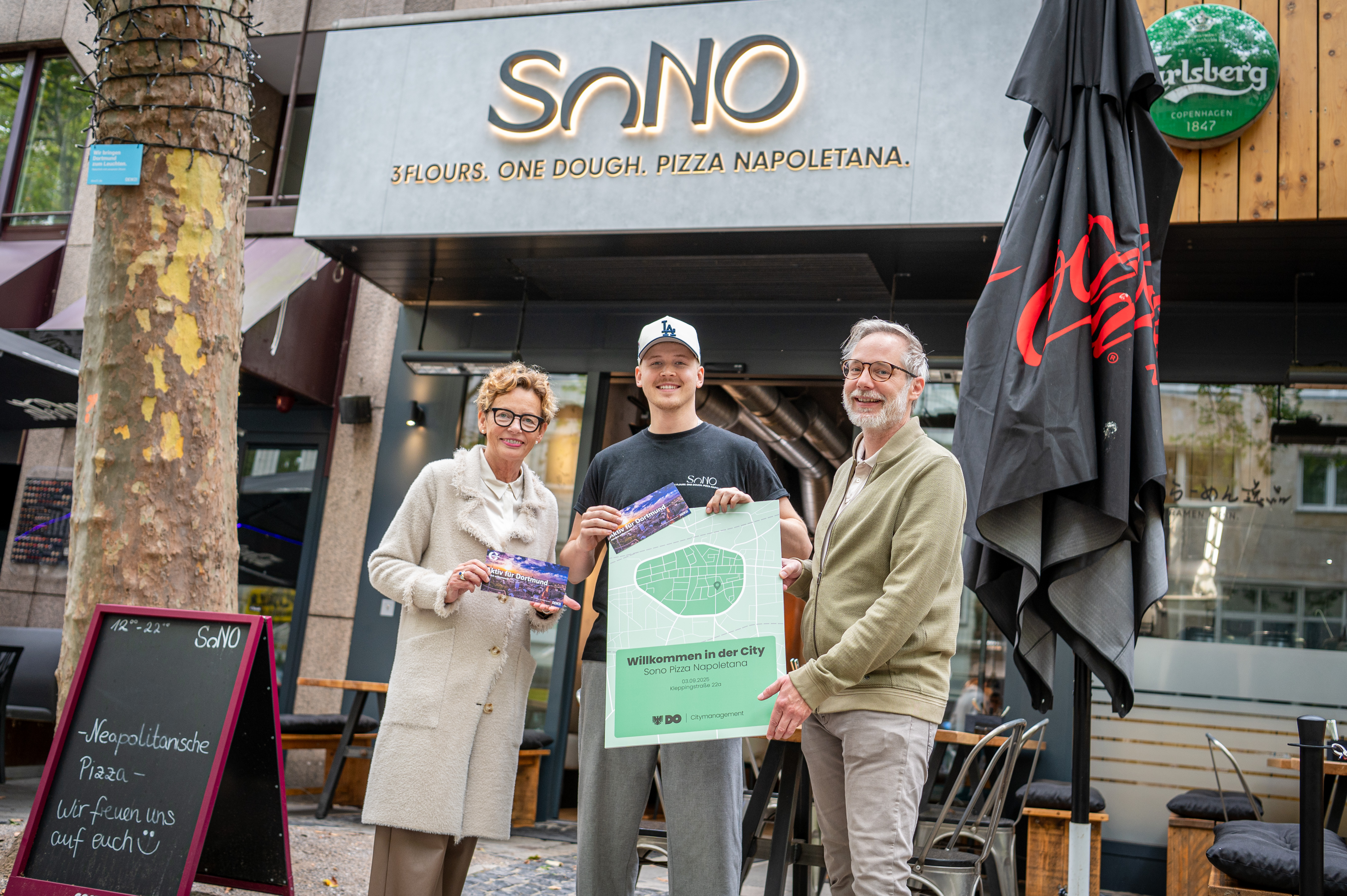 Zwei Männer und eine Frau stehen vor einer Pizzeria und halten eine stilisierte Karte der Dortmunder City.