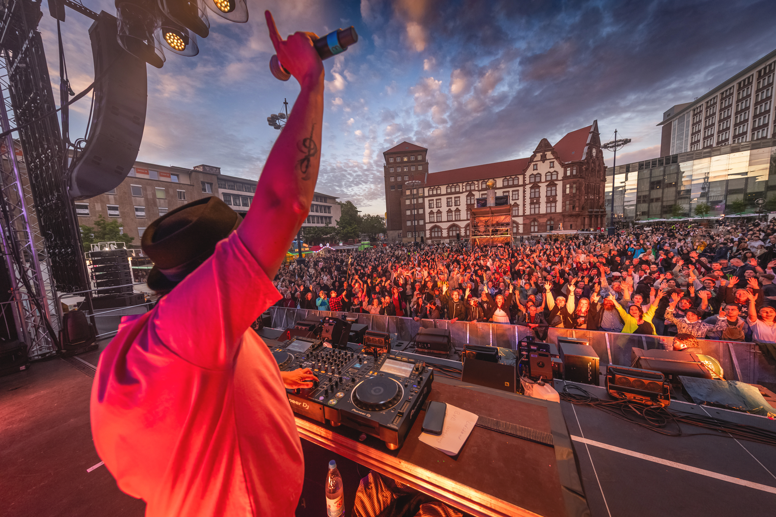 Ein DJ, der auf einer Bühne eine Hand hochreckt, im Hintergrund eine Menschenmenge.