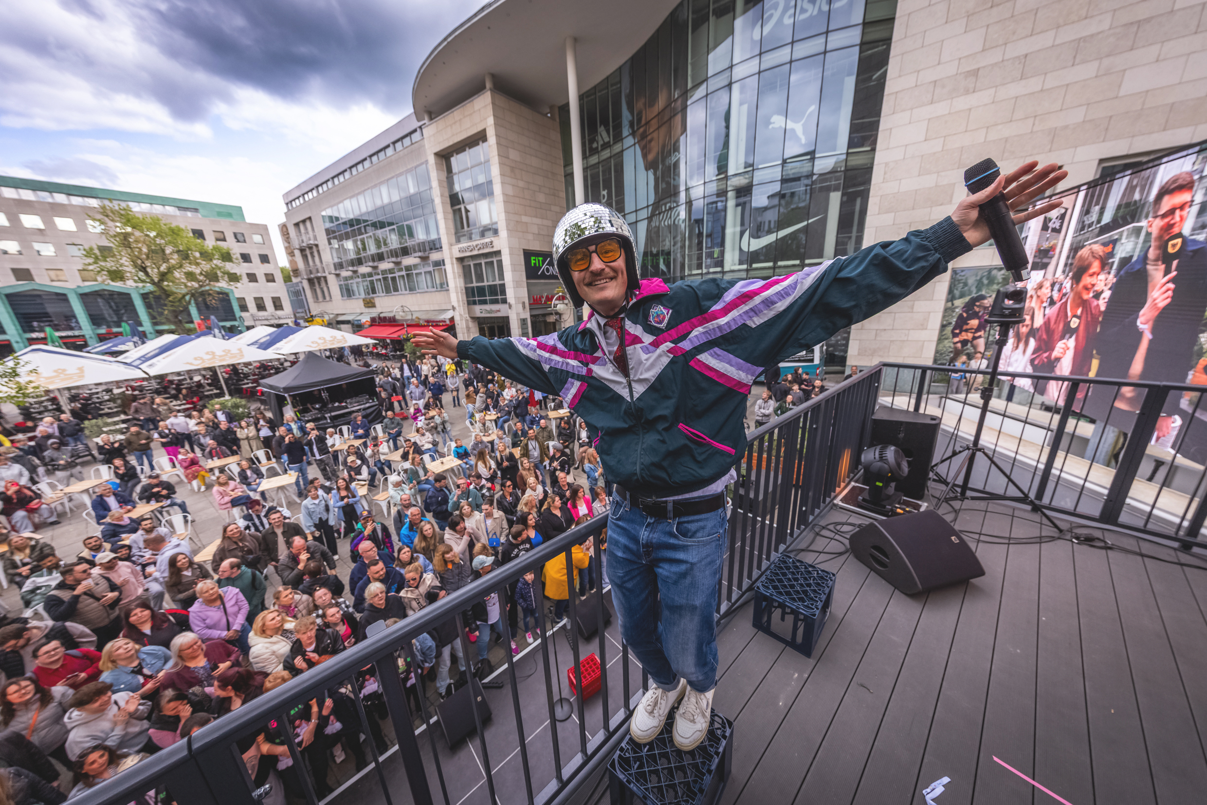 Foto eines Mannes mit einem Discokugel-Helm auf einer hohen Bühne vor einer Menschenmenge. 
