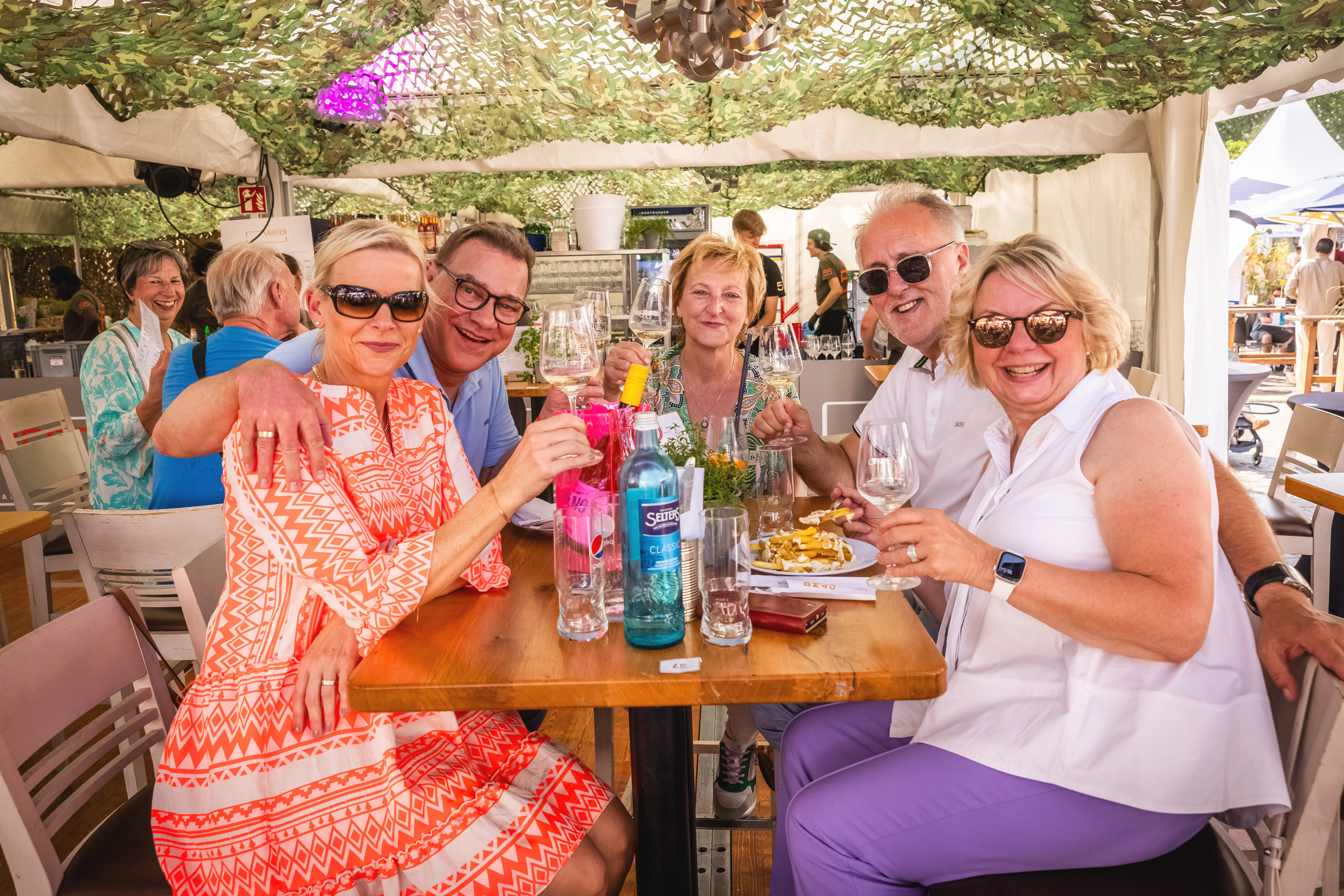 Eine Gruppe von Menschen, die bei Sonnenschein in einem weißen Zelt essen und trinken und in die Kamera schauen.