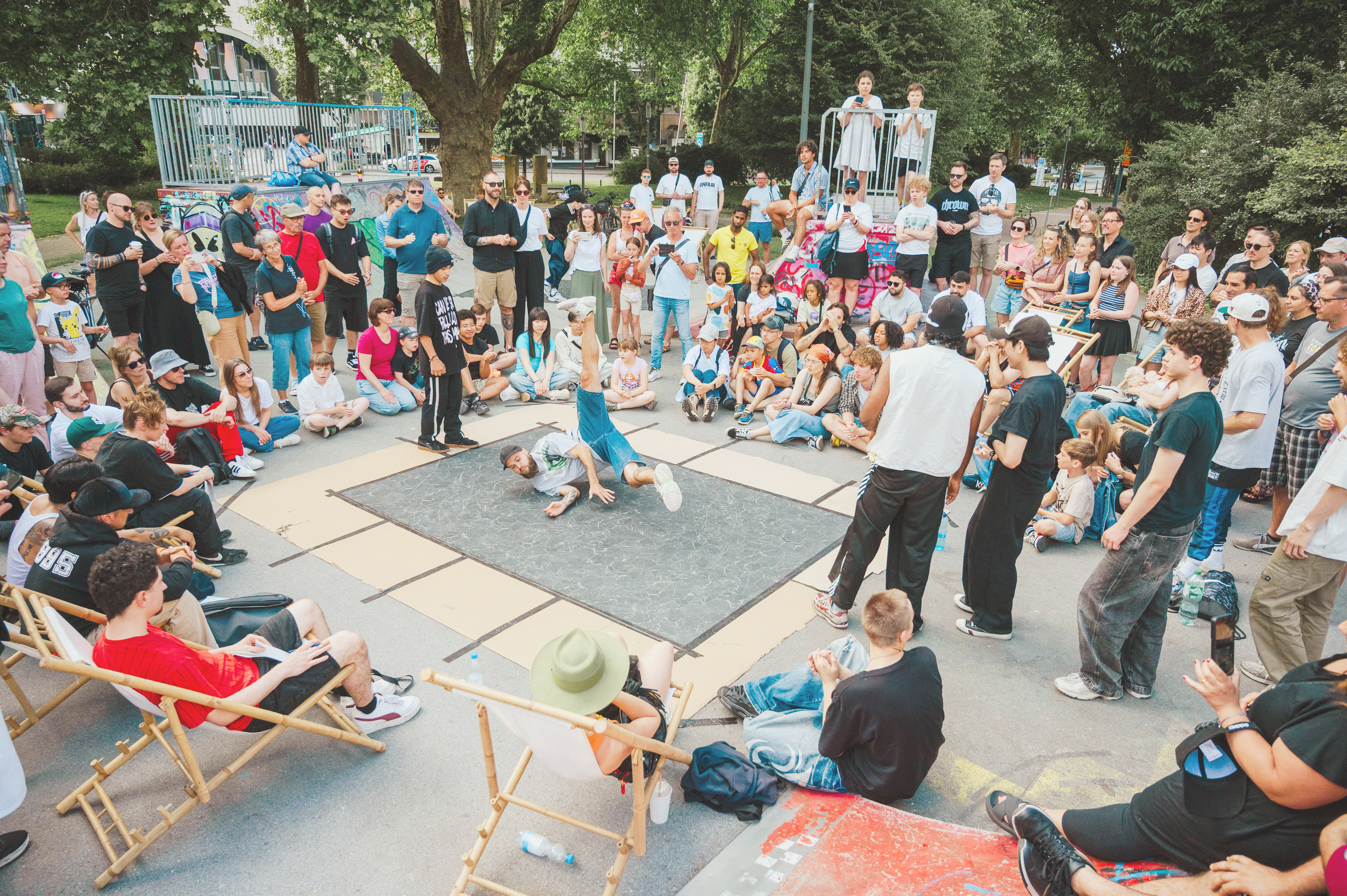 Ein Breakers auf einer Matte, umringt von Menschen, im Hintergrund eine Skate-Anlage.