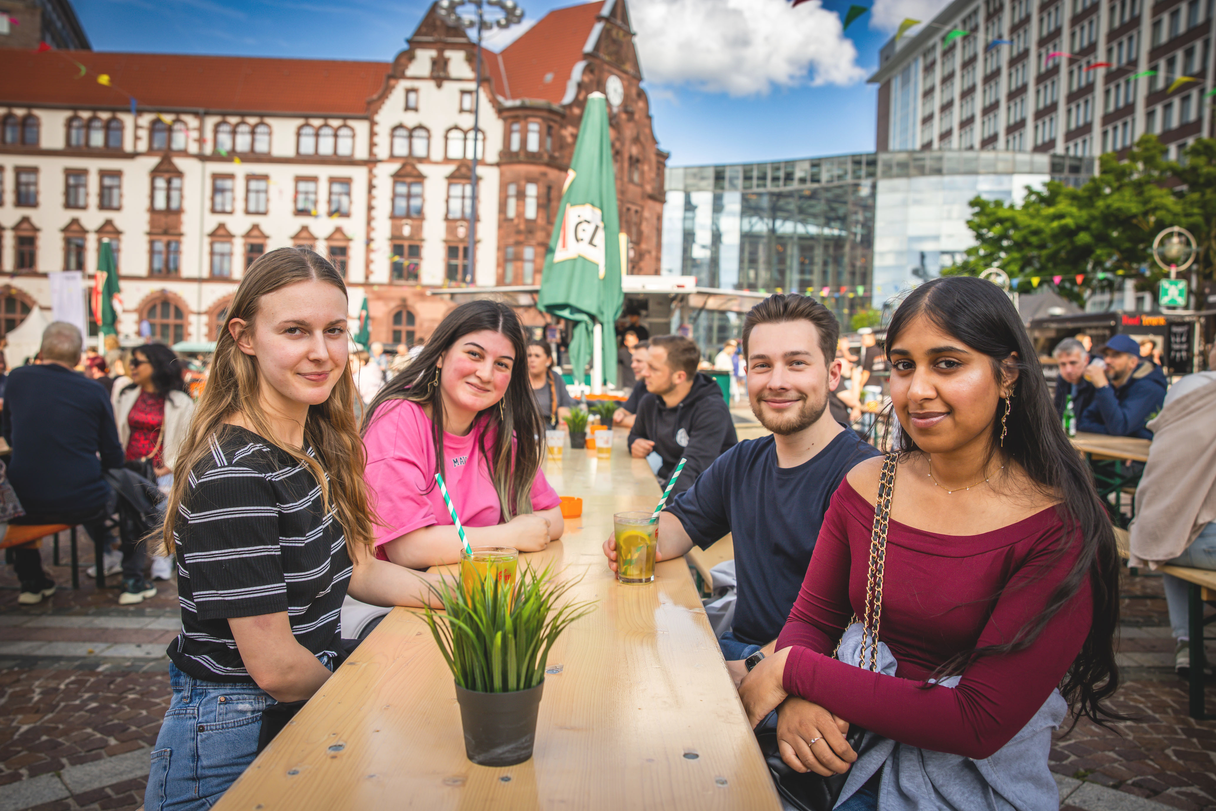 Vier junge Erwachsene sitzen an einer Bierzeltgarnitur auf dem Friedensplatz, im Hintergrund das Alte Rathaus.
