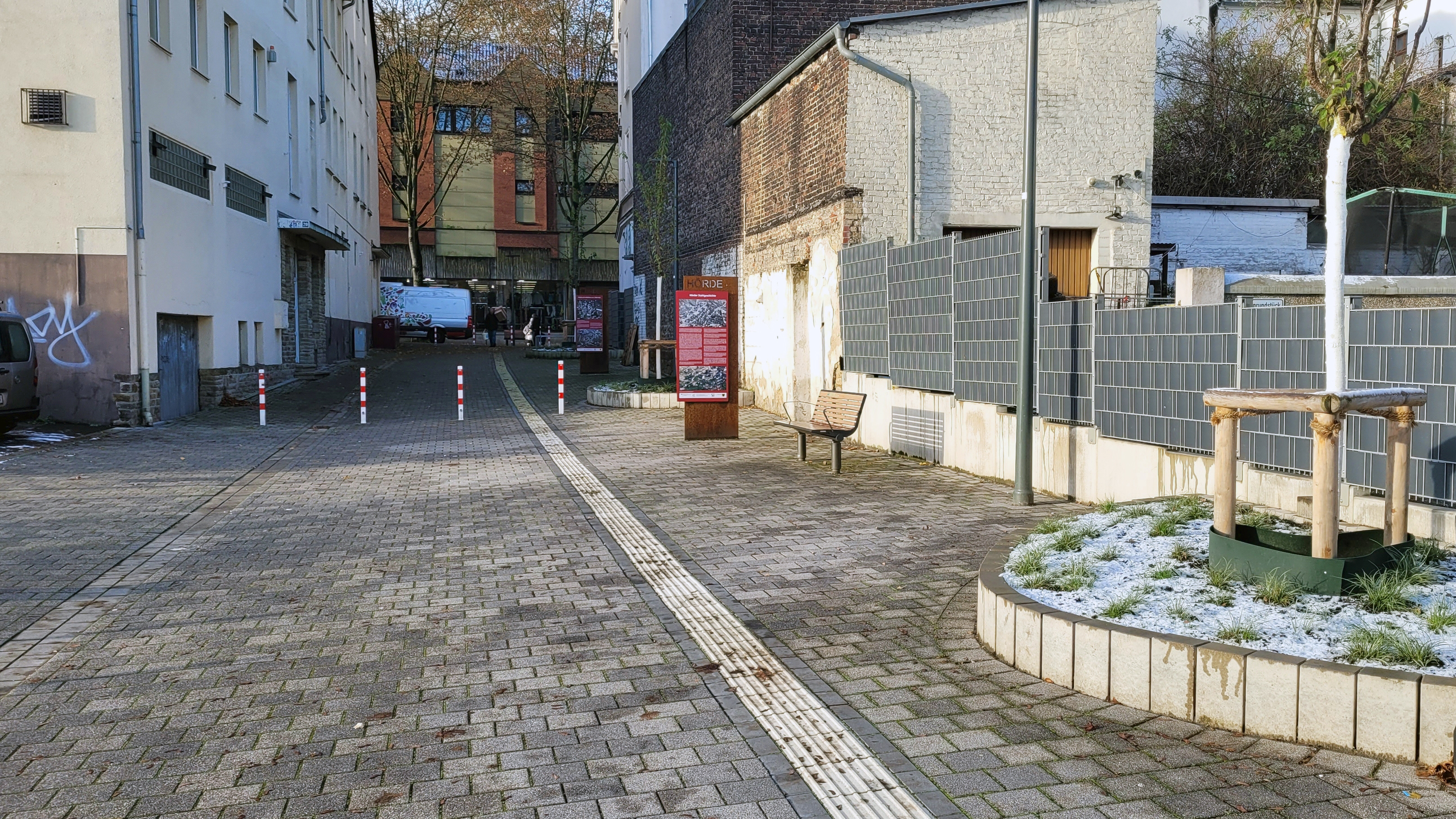 Foto einer frisch gepflasterten Fläche mit neugeopflanztem Baum, taktilem Leitsystem und einer Infostele.