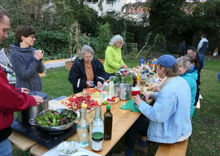 Mehrere Menschen stehen in einem Garten an einem langen Tisch und bereiten Gemüse zum Kochen vor.