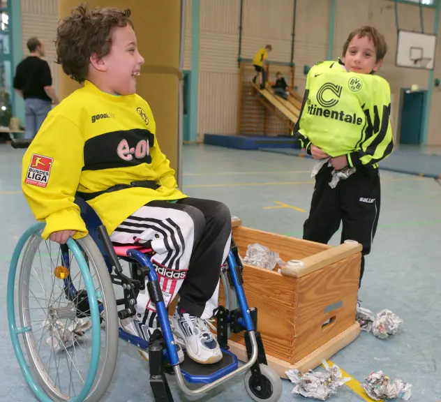 Zwei Jungen mit BvB-Trikot stehen in einer Turnhalle und lachen, einer der Jungen sitzt in einem Rollstuhl