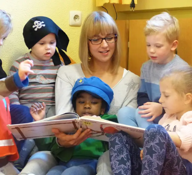 Eine Frau mit blonden Haaren und Brille liest sechs Kindern in Verkleidung ein Buch vor