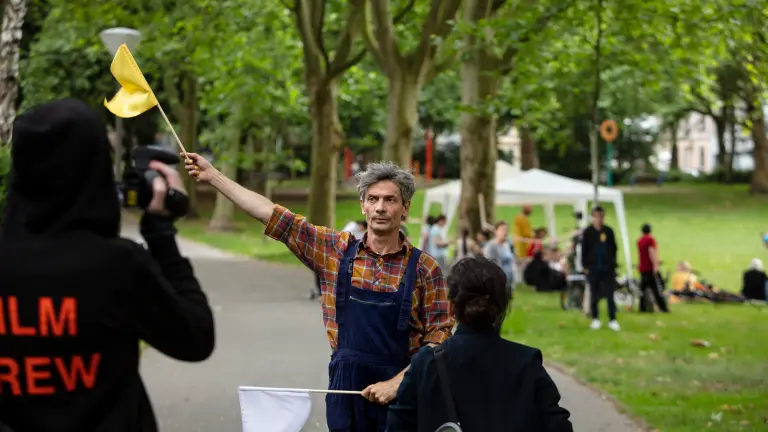 Eine Filmcrew filmt in einem Park einen Mann mit grauem Haar, kariertem Hemd, Latzhose und einer kleinen gelben Flagge, die er in die Luft hält