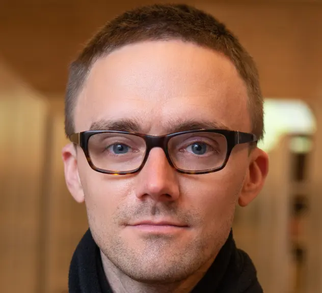 Portrait eines Mannes mit kurzen braunen Haaren, schwarzer Brille und schwarzem Schal 