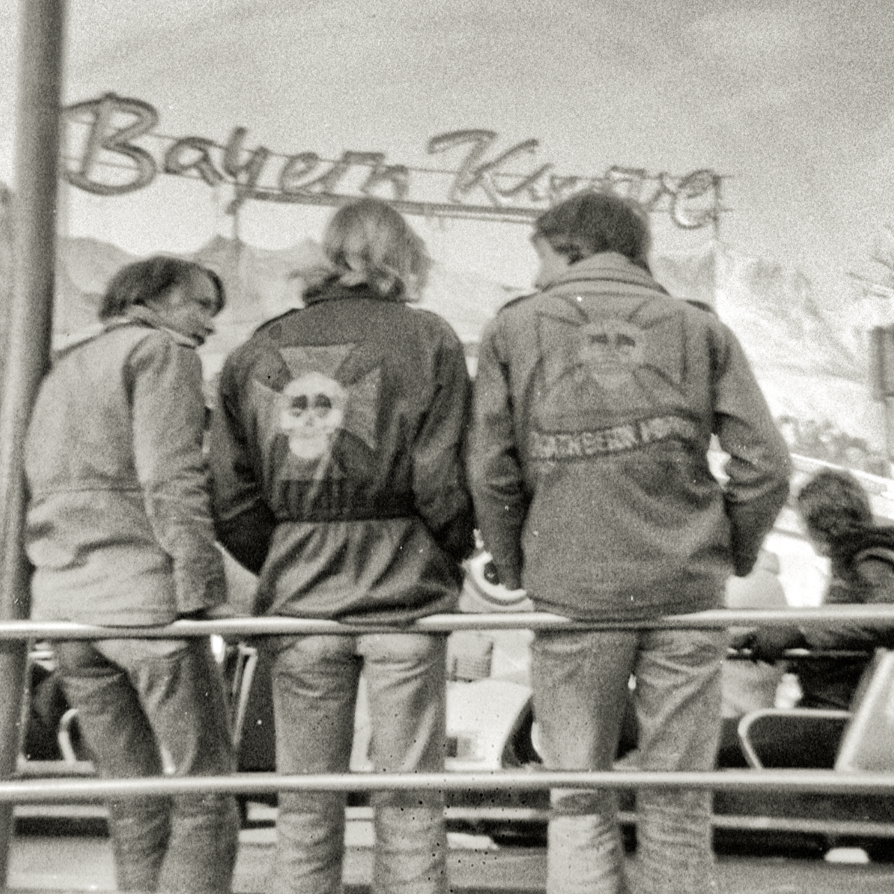 Schwarz-weiß Foto von drei Jugendlichen, die von hinten zu sehen sind. Sie stehen vor einer Achterbahn.