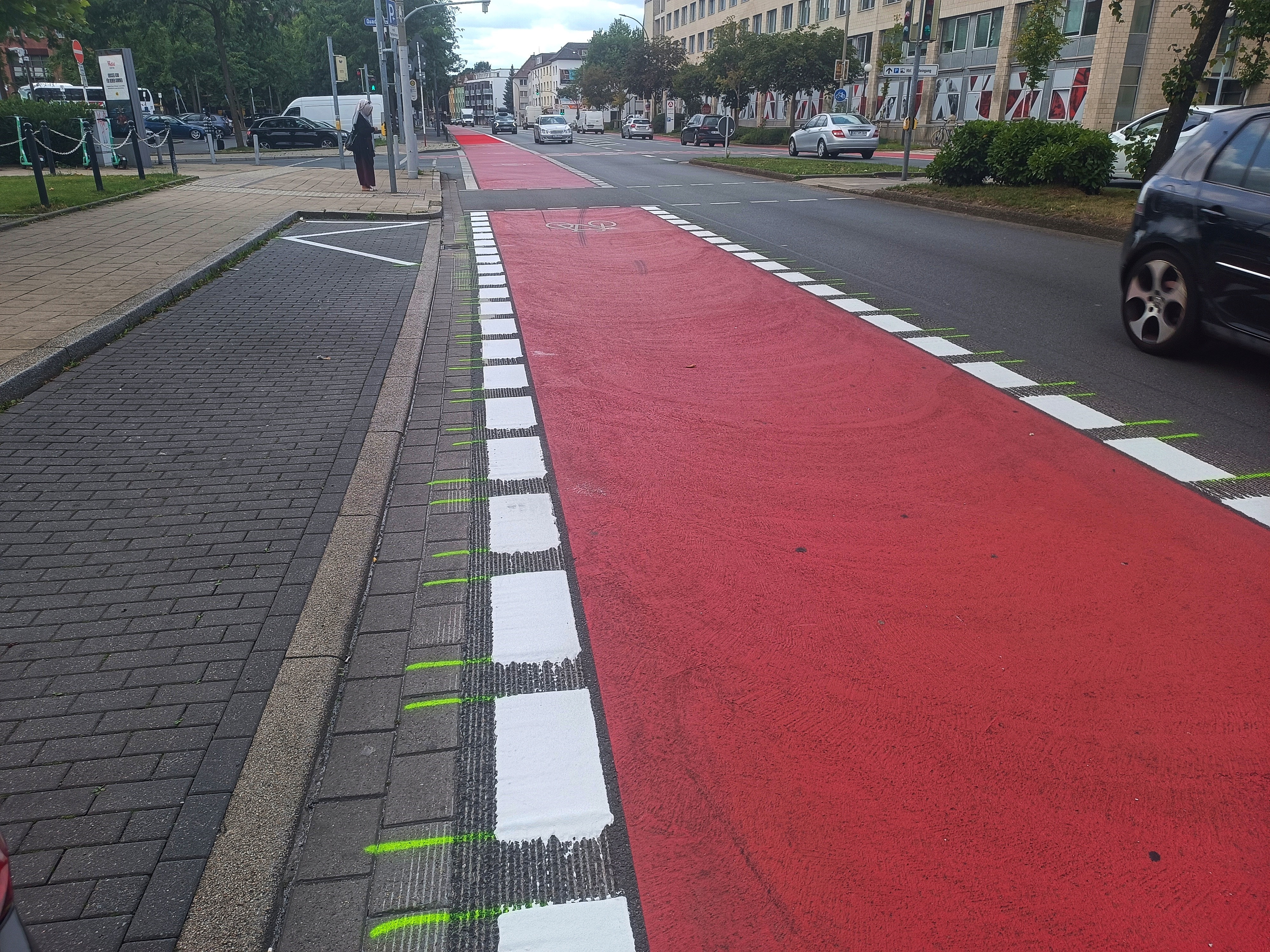 Ein roter Radfahrstreifen ist zu sehen. Die beiden weißen Leitlinien sind links und recht unterbrochen. Im rechten Bildrand fährt ein Auto. Links ist eine Haltebucht zu sehen. 